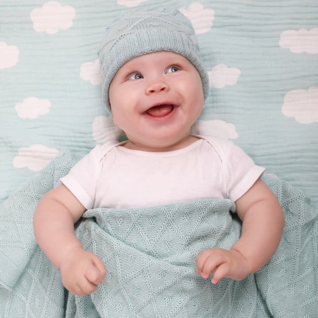 petit bébé qui regarde en haut et sourit sur son lit portant un bonnet et couvert d'une housse