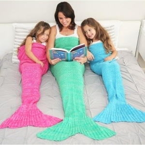 Une maman dans le lit avec ces deux filles qui sont avec la couverture en forme de sirène une rose une bleue et une verte