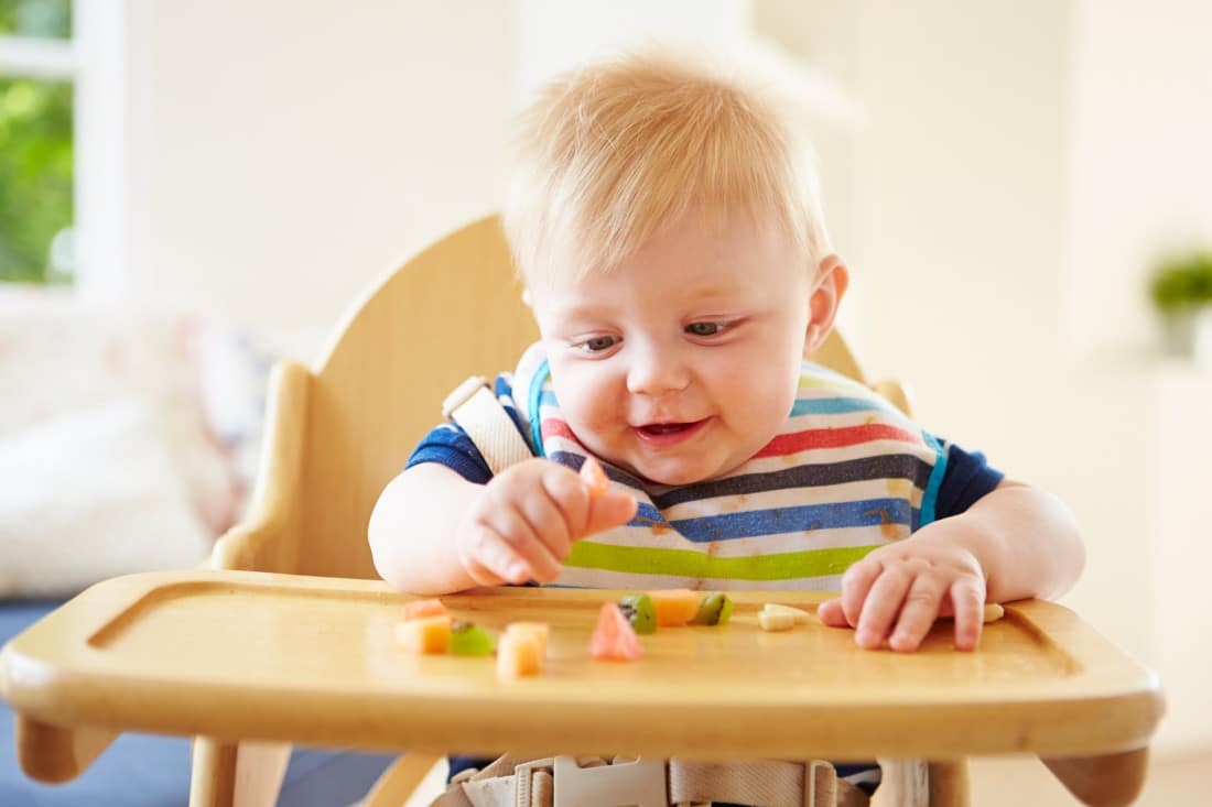un bébé assis dans sa chaise haute qui mange des fruits coupés en petits morceaux 