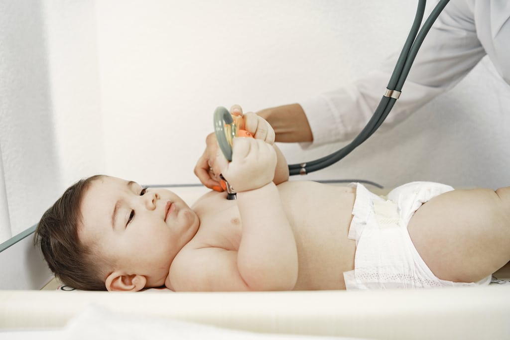 un bébé allongé chez le pédiatre tient un jouet entre ses mains pendant que le pédiatre pose le stéthoscope sur sa poitrine