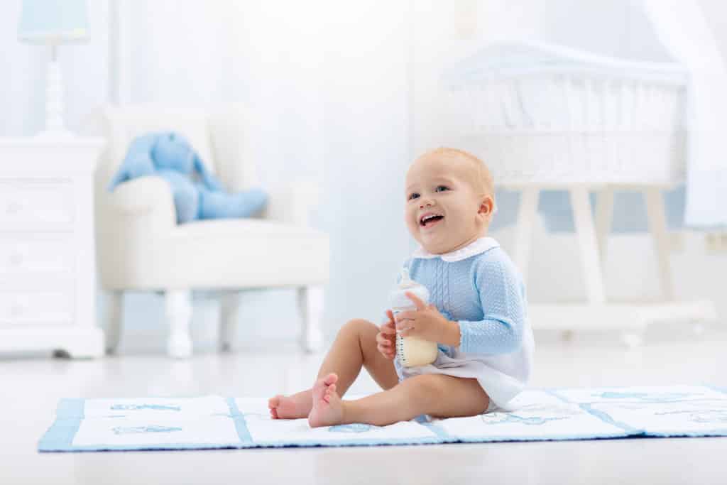 un bébé assis par terre sur une serviette bleue fait un grand sourire tandis qu'il tient un biberon de lait dans ses mains