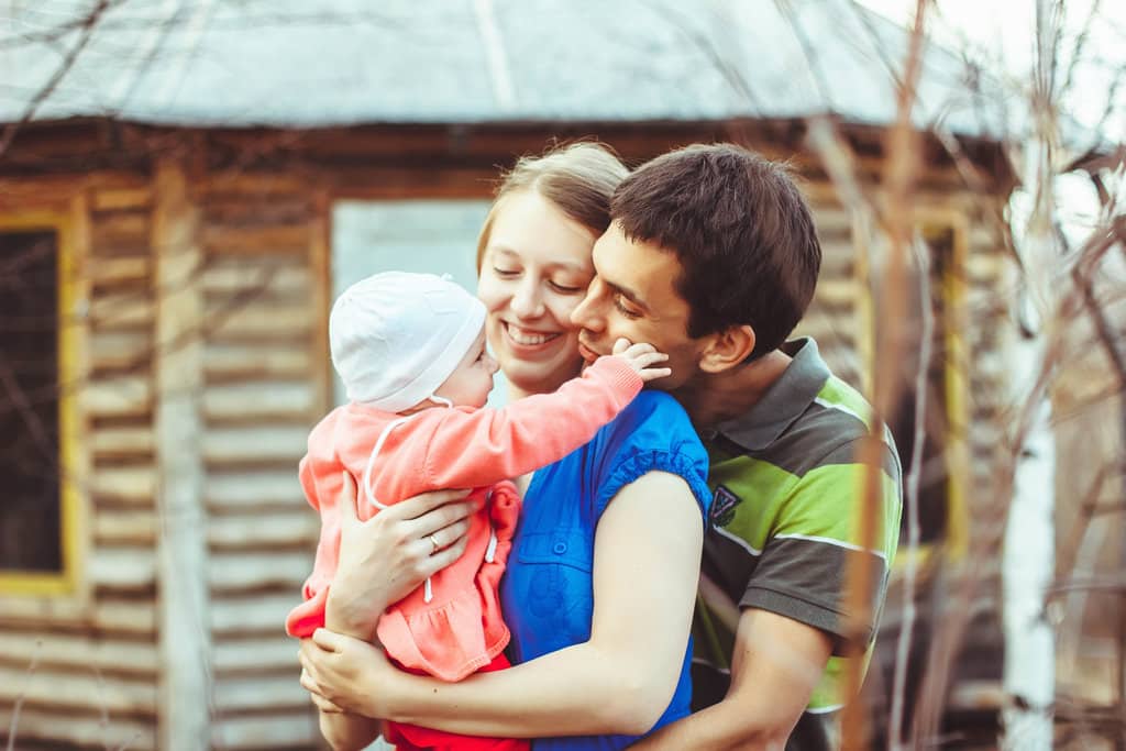 Devant un chalet en bois, 2 parents et leur bébé en premier plan. Le papa entoure la maman avec ses bras et approche son visage du bébé qui est porté dans les bras de sa mère.