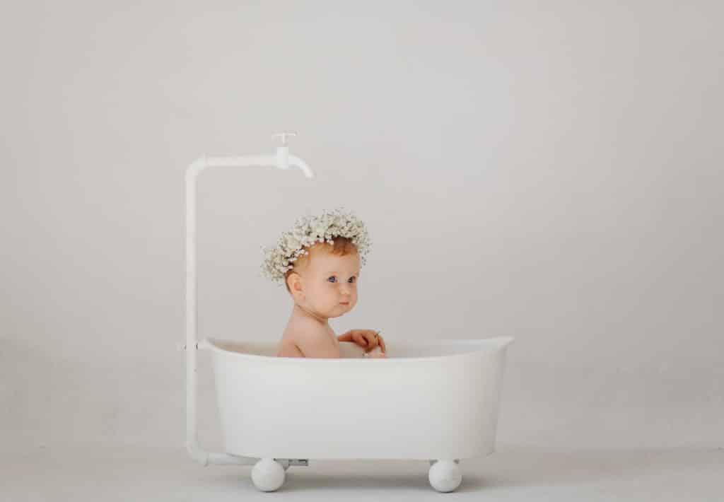 photo issue d'un shooting bébé. un bébé avec une couronne de fleurs blanches sur la tête, est assis dans une baignoire toute blanche à roulettes. il a un robinet blanc au-dessus de la tête
