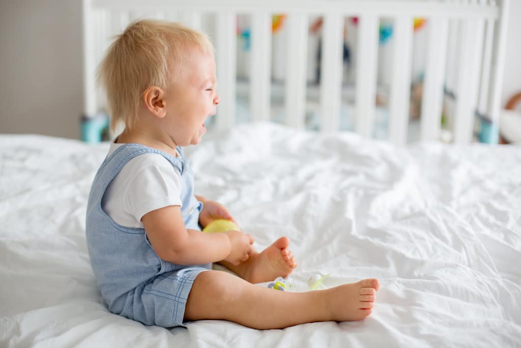 un bébé en body blanc et salopette bleue, est assis sur un grand lit blanc et pleure tout seul
