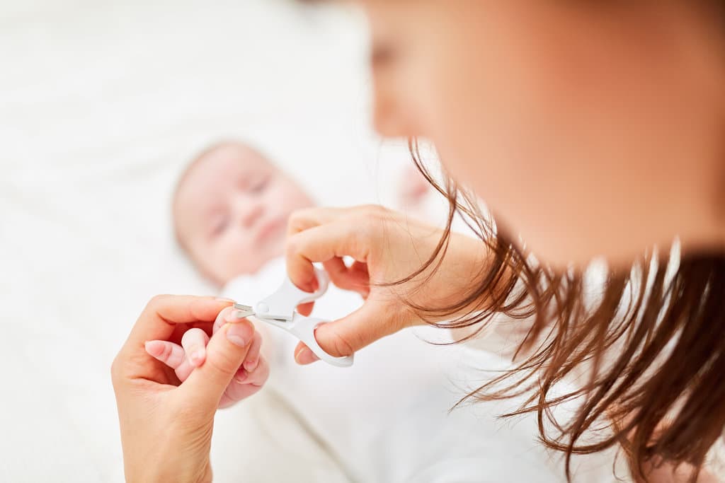 un bébé allongé a la main tendu tenue par sa mère qui lui coupe les ongles avec un ciseau à bouts ronds