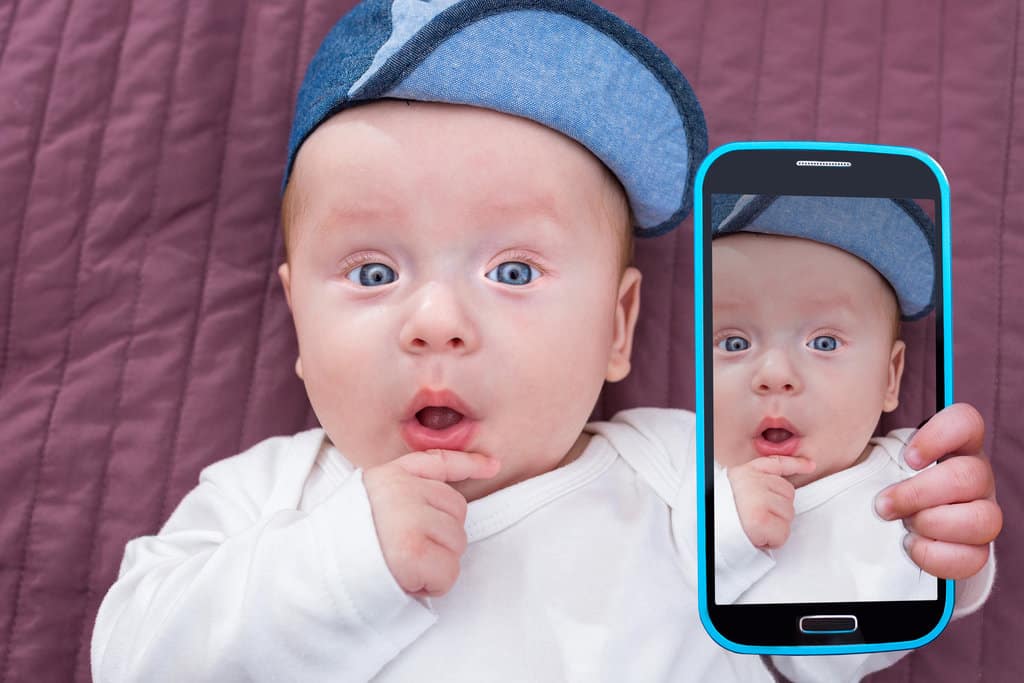 un bébé est allongé sur une couverture violette qui sert de fond pour la photo. il porte une casquette grise, a la bouche ouverte, les yeux bleus grands ouverts et met sa main droit sur son menton. Sa main gauche tient un smartphone en train de le prendre en photo. Il fait un selfie