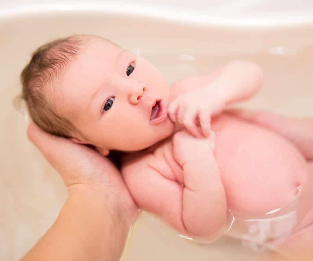 le meilleur moyen de donner le bain à bébé est dans sa baignoire. la bouche ouverte et les bras contre la poitrine. une main adulte lui tient la tête et la nuqye