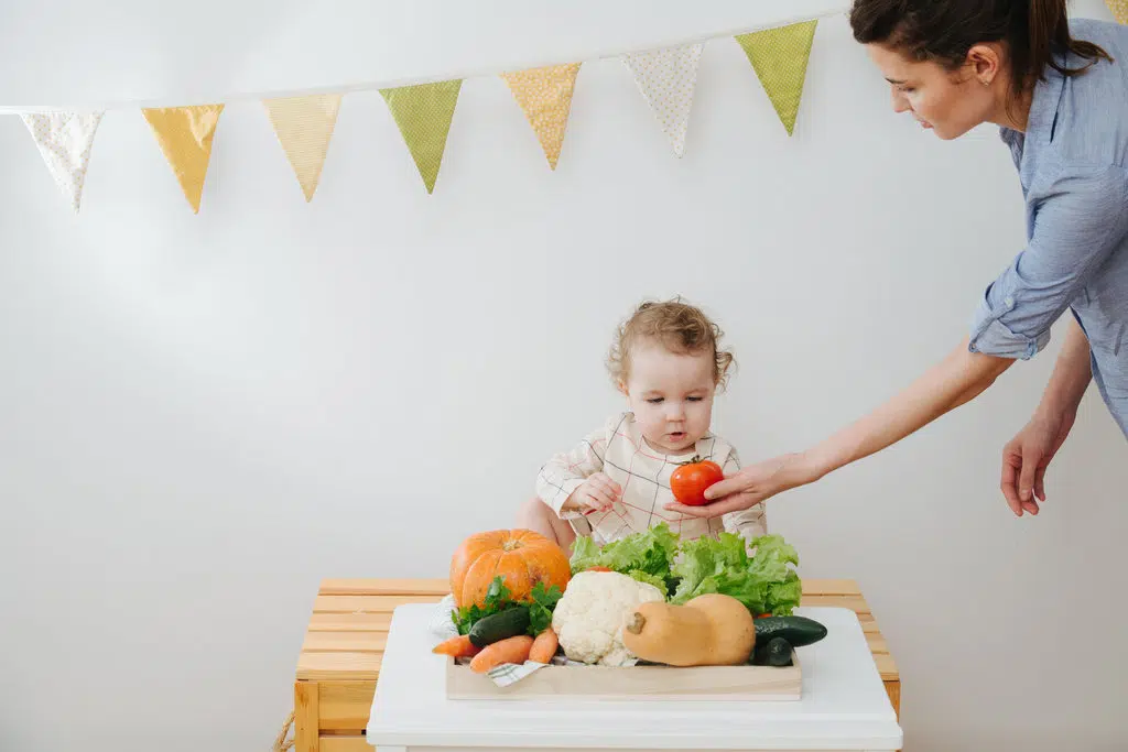un bébé est devant une table sur laquelle sont disposés des légumes frais et regarde avec étonnement une tomate que sa mère lui met devant son visage