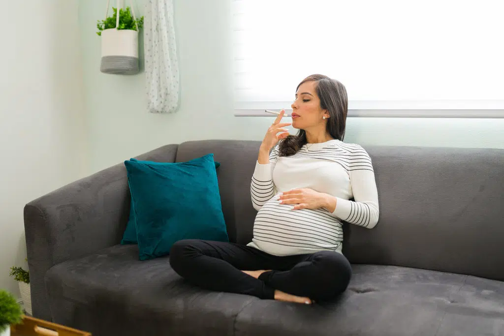 une femme enceinte fume une cigarette, assise en tailleur sur un canapé gris et prend le risque d'avoir un bébé qui tousse