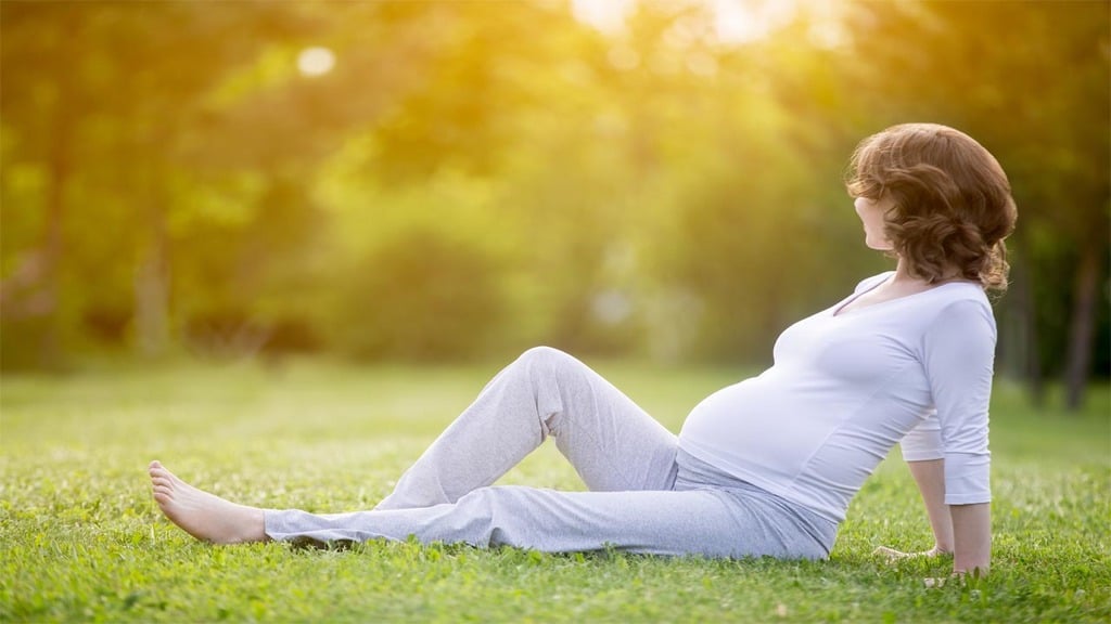 une femme enceinte de 5 mois, vêtue de blanc, se prélasse dans l'herbe d'un parc arboré