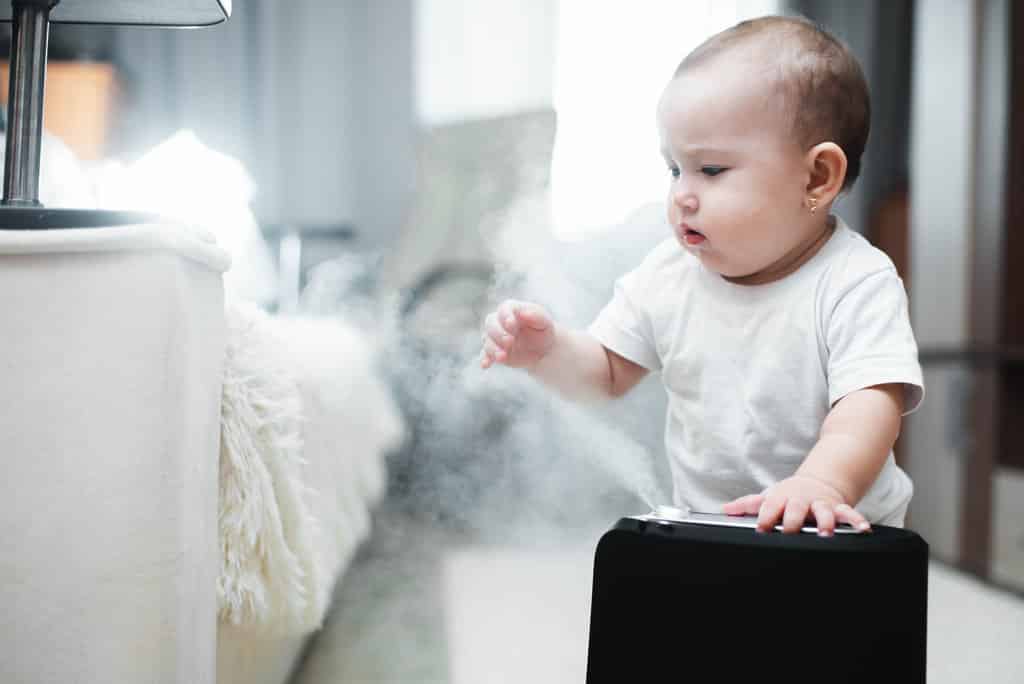 un bébé en body blanc manches courtes joue avec un humidificateur d'air à vapeur dans une chambre près d'un lit