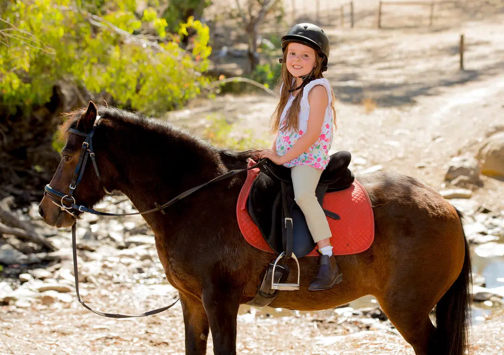 une petite fille est sur un poney avec son casque de sécurité, prête pour partir en promenade