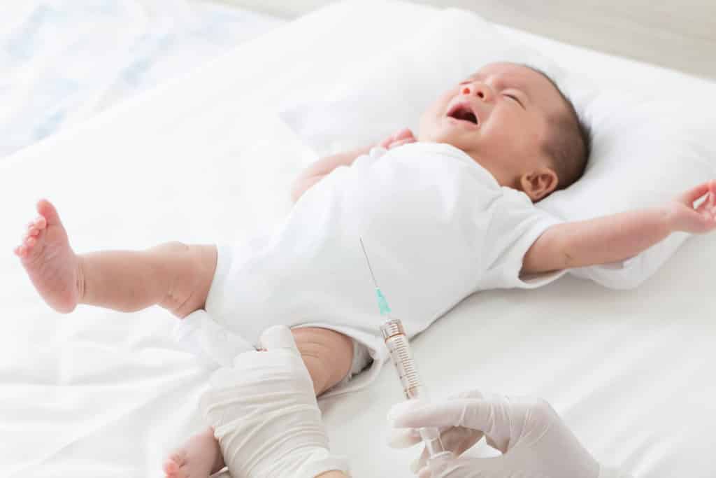 un bébé allongé sur un lit médical, pleure. on y voit 2 mains gantés dont l'une retient la jambe du bébé et l'autre tient la seringue  contenant le vaccin