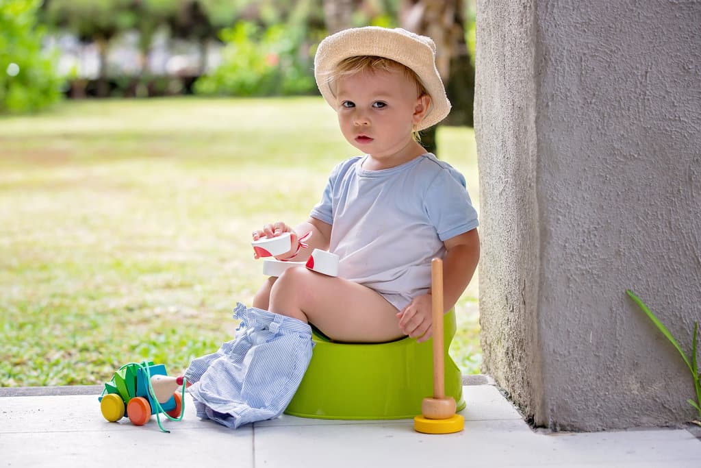 un bébé est assis sur le pot sur la terrasse dehors. il porte un chapeau et un tee-shirt. Son pantalon est détroussé sur ses pieds. A côté de lui et dans ses mains, il y a des jouets pour l'occuper.