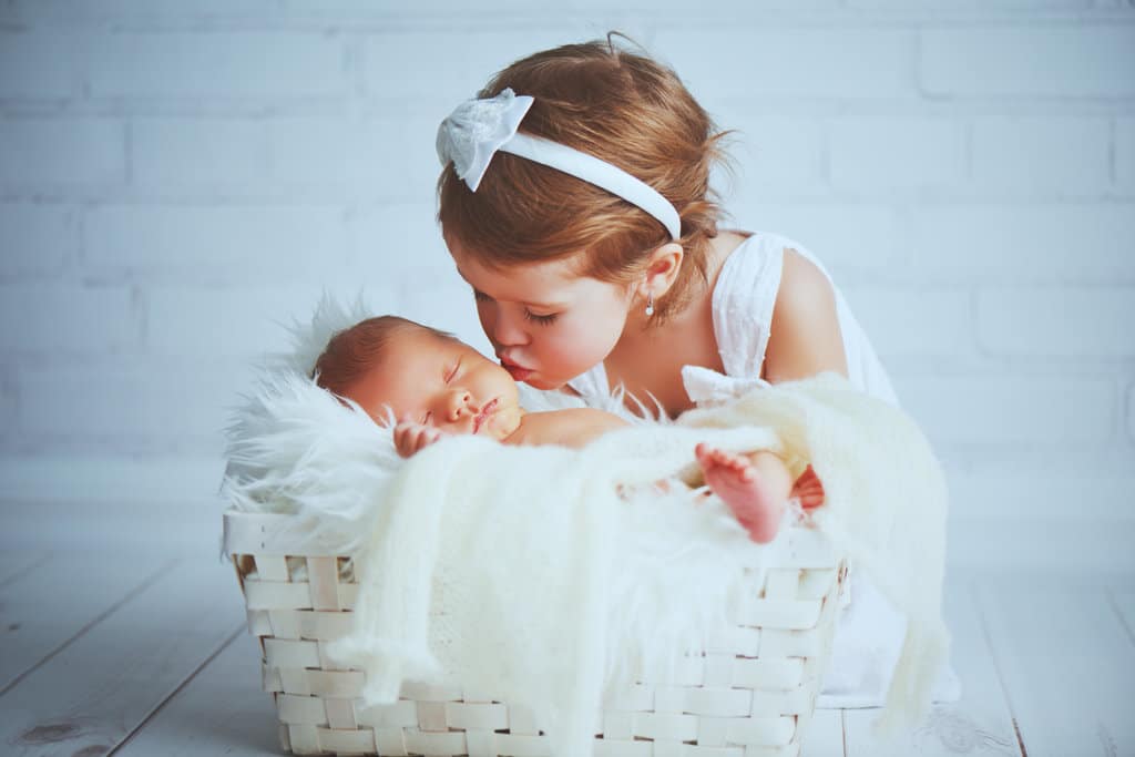 lors d'une séance photo bébé, un nouveau-né est endormi dans un panier sur une couverture à long poils blancs. sa petite soeur habillée en robe blanche et serre-tête blanc, lui fait un bisou sur la joue