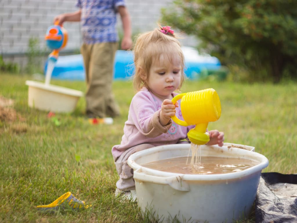 une petite fille fait une démonstration de comment occuper un bébé de 18 mois. Elle tient un arrosoir jaune dans sa main, qui déverse de l'eau dans une grande bassine d'eau marron. tout cela se passe dans le jardin, avec en fond un garçon plus grand qui fait la même activité avec une autre bassine et un autre arrosoir