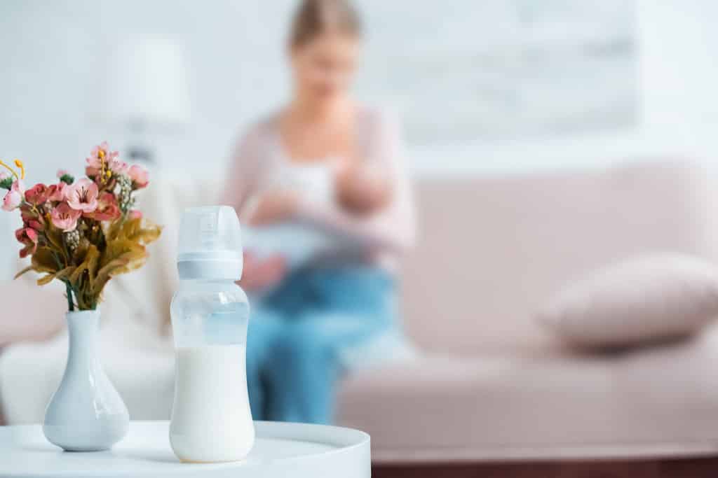 Vue rapprochée sur un biberon de lait et des fleurs dans un vase. Une mère allaite son bébé dans le fond, posée sur un canapé