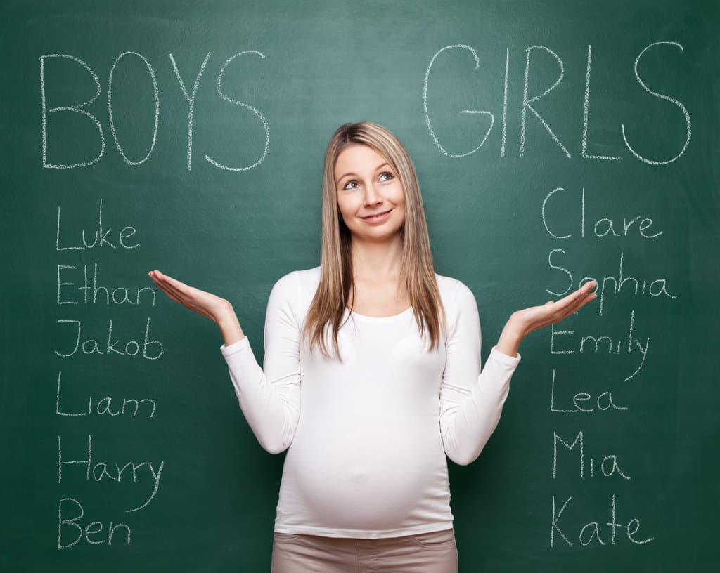 une femme enceinte a écrit à la craie sur un tableau un liste de prénoms de garçon et une liste de prénom de filles. les paumes vers le ciel, elle se demande bien quel prénom choisir