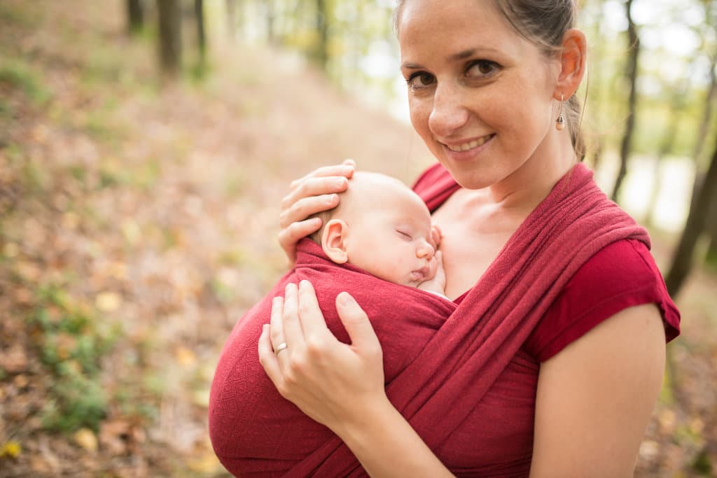 dans une forêt, une maman prend la pose avec son nouveau-né dans une écharpe de portage rouge