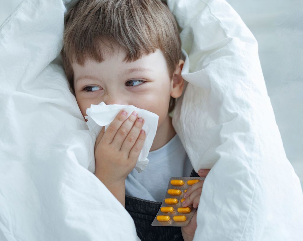 un enfant enveloppé dans une couette blanche porte à son nez un mouchoir et tient dans l'autre main une plaquette de médicaments