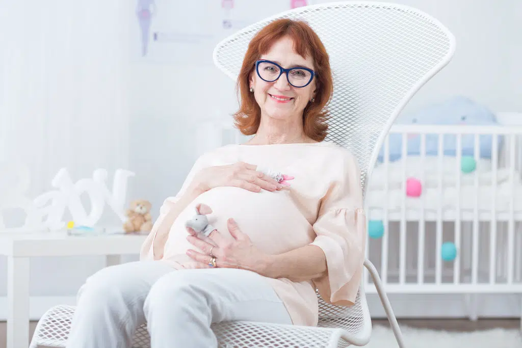une femme enceinte d'un certain âge portant des lunettes bleues, est assise sur une chaise dans un chambre de bébé.