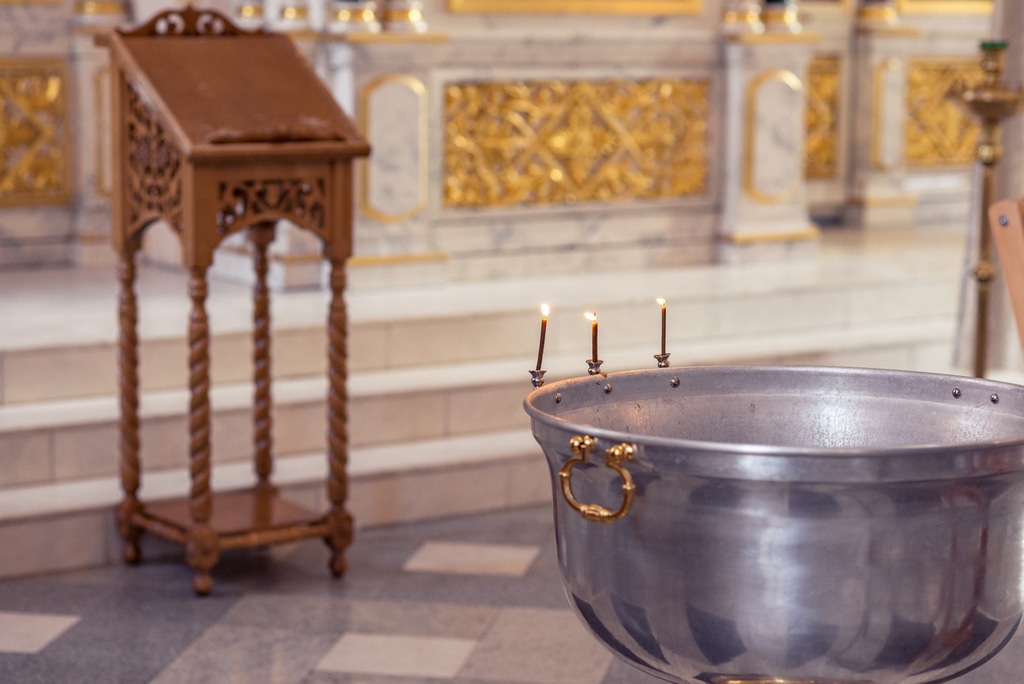 on aperçoit au premier plan, dans une église, un bénitier avec 3 petites bougies allumées. au fond, les marches de l'autel ainsi qu'un pupitre en bois.