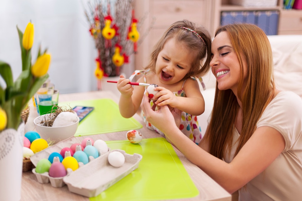 une femme aide une petite fille à peindre un oeuf en lui tenant l'oeuf avec sa main. Tout sourire, la petite fille tient un pinceau et peint l'oeuf tendu par la femme. Devant elles, sur une table, se trouve un boite d'oeufs peints.