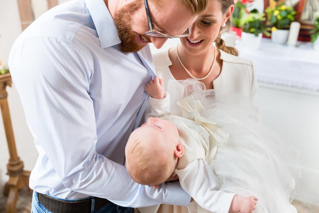 les parents tiennent leur bébé fille habillée d'une jolie robe blanche pour la faire baptiser. elle en profite pour tirer sur la chemise de son papa.