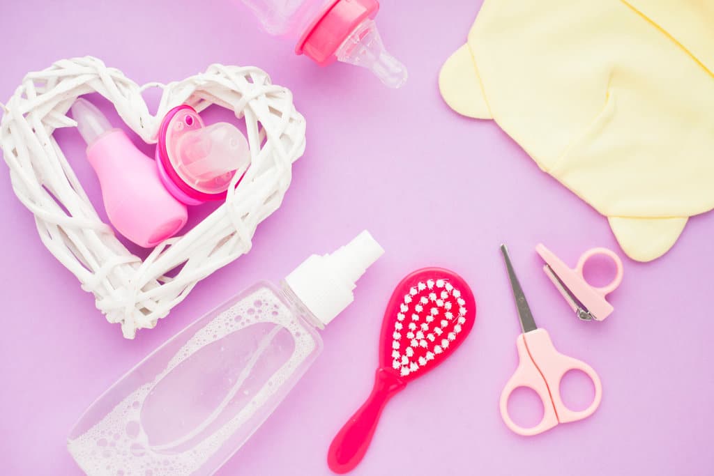 divers accessoires de toilette bébé roses sont posés sur un fond violet. on y voit une brosse, un ciseaux à bout rond, une pince à ongles, une bouteille, un mouche bébé, une sucette, une tétine.