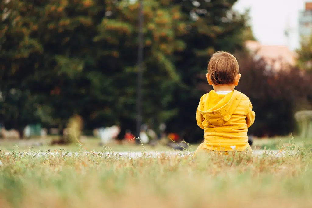 un enfant avec un sweat capuche jaune est assis, de dos, seul dans l'herbe
