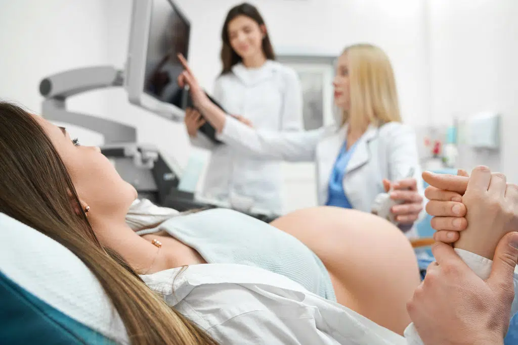 une femme enceinte est allongée dans un cabinet médical et tient la main de son conjoint pendant qu'une gynécologue et son assistante regardent son échographie