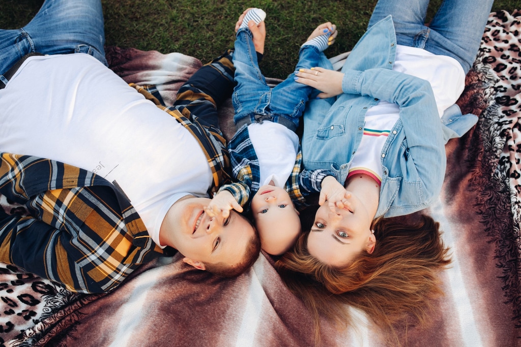 photo prise vue du dessus sur une couple allongé dans l'herbe avec leur enfant au milieu d'eux. ils portent tous un jeans et une chemise ouverte sur un t-shirt. les parents tiennent chacun un pied de l'enfant qui écarte les bras en direction de la photo