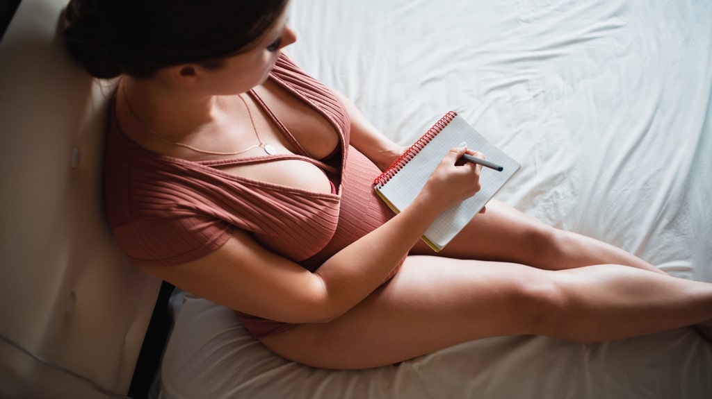 vue du dessus sur une femme enceinte avec un décolleté qui écrit la liste de naissance sur un calepin. elle est assise sur un lit