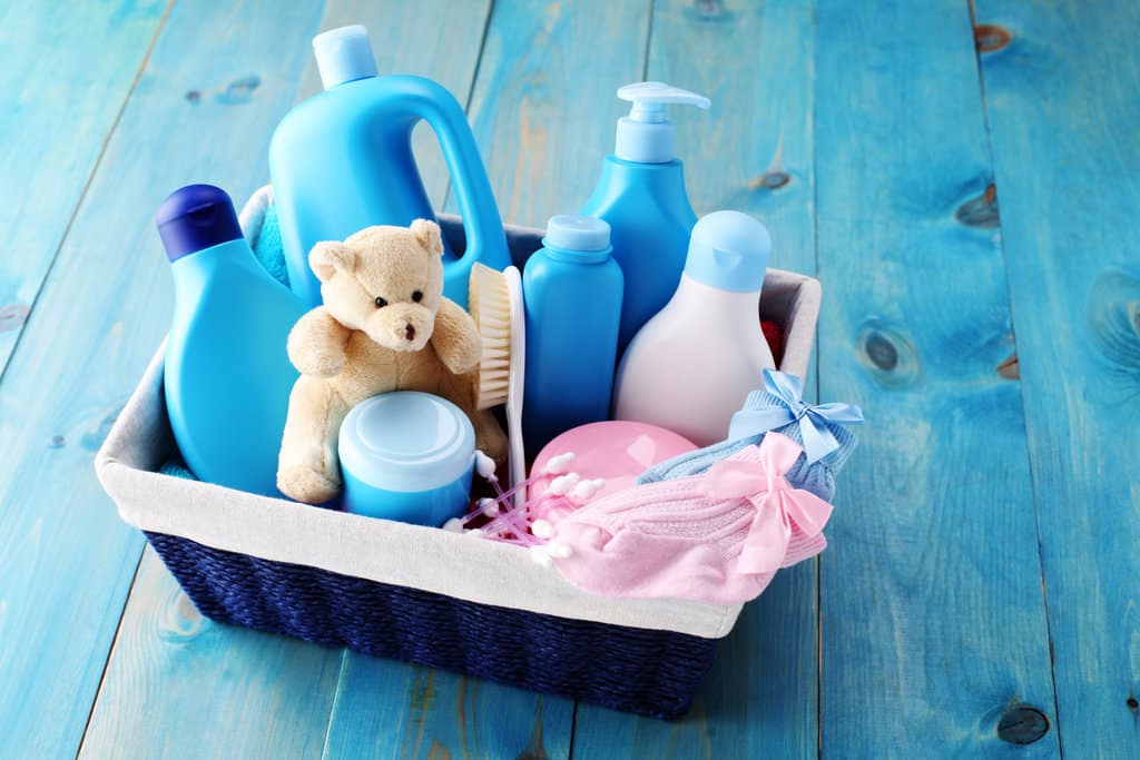 un panier bleu est posé sur un sol en bois bleu. dans ce panier contient des bouteilles de savon sans étiquette, un petit ours, des cotons tiges et des petites chaussettes bébé : une rose et une bleue.