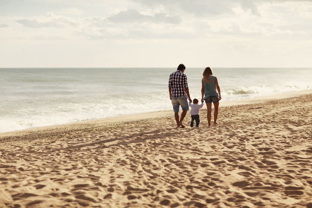 sur une plage de sable, des parents se baladent en tenant la main de leur bébé qui marche entre eux deux.