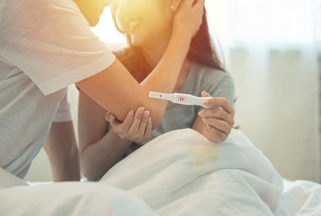 un couple est dans leur lit, assis, et se regardent tendrement. la femme tient un test de grossesse positif qu'elle met en avant sur la photo.