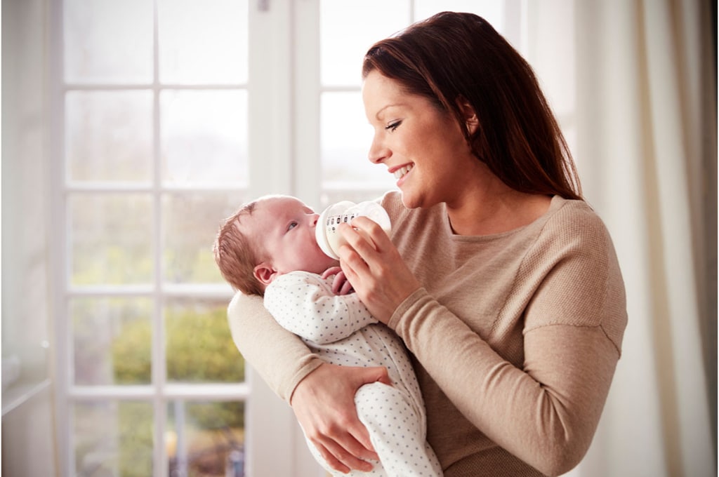 une maman qui se tient debout donne le biberon à son bébé qu'elle tient au creux de son bras. elle porte une robe beige et sourit à son bébé qui la regarde.