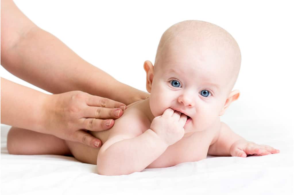 un bébé couché sur le ventre avec des yeux bleus, a des mains posées sur son dos. il porte 3 doigts de sa main dans la bouche