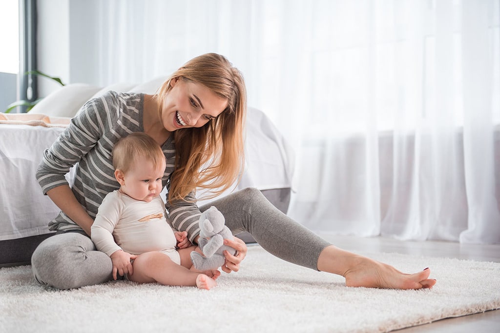une mère est assise parterre sur un tapis blanc avec son bébé assis contre elle. elle sourit tout en lui montrant un nounours gris devant lui.