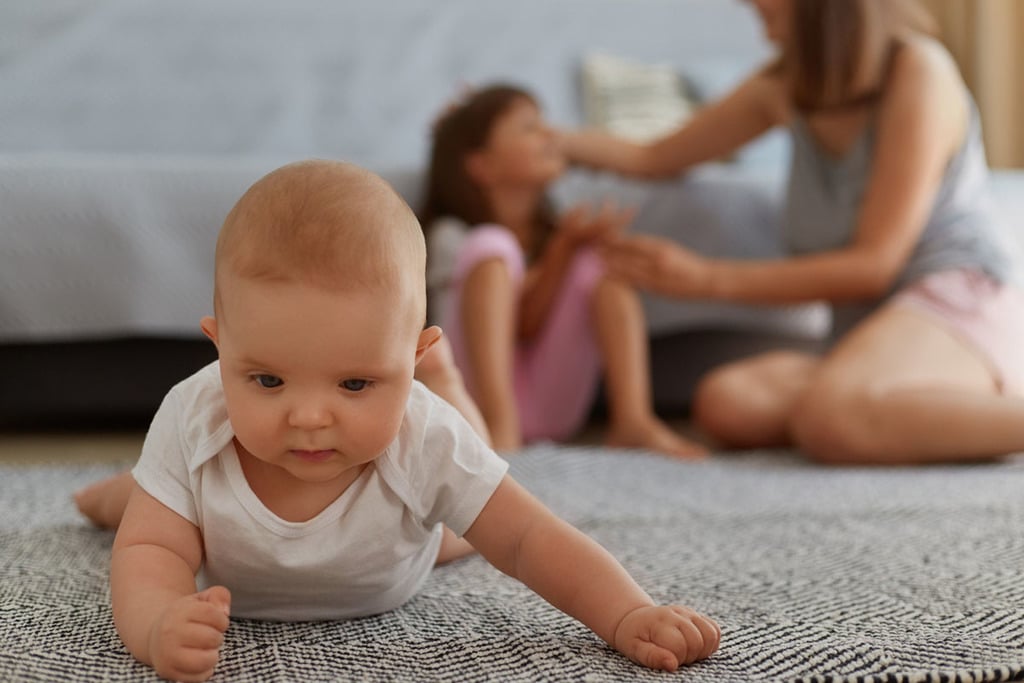 un bébé est à plat ventre sur un tapis dans le salon, avec la tête relevée et qui se tient à l'aide de ses bras. dans le fond, on aperçoit une jeune fille et sa mère qui discutent assises sur le tapis.