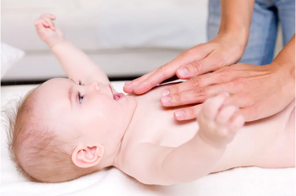 un bébé sur le dos a les bras écartés tout heureux de se faire masser par des mains d'adulte posées au-dessus de son ventre