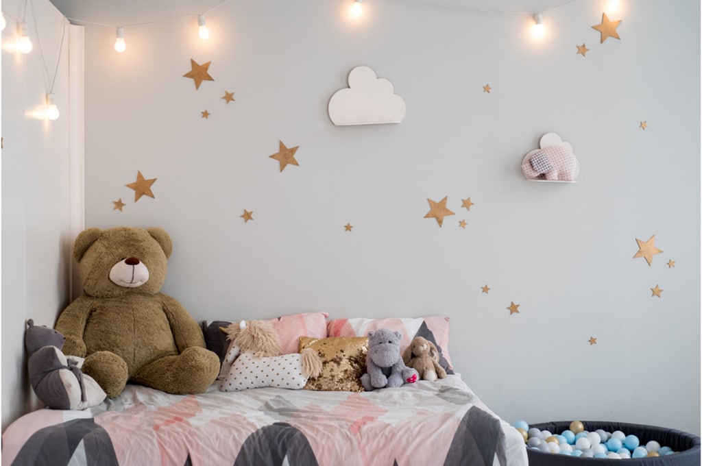 chambre d'enfant avec des murs blancs sur lesquels sont accrochées des étoiles et des nuages. plusieurs peluches dont un gros nounours sont posés sur le lit. à droite, il y a une piscine à balles.