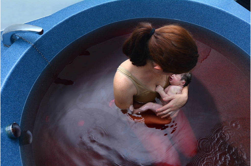 vue du dessus sur une baignoire remplie d'eau et de sang, dans laquelle une femme vient d'accoucher. Elle est agenouillée avec son nouveau-né dans les bras qu'elle regarde.