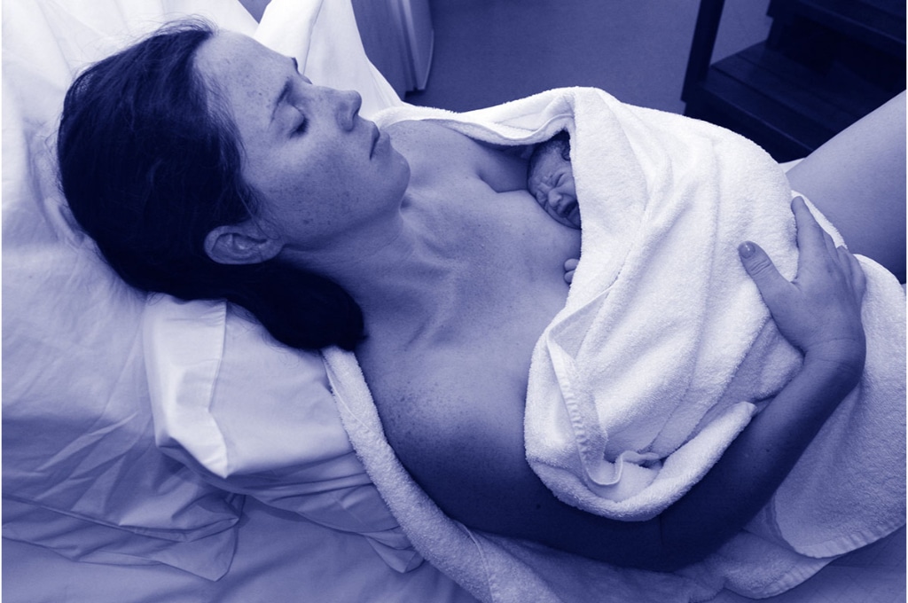 une femme est nue entourée d'une serviette blanche avec son nouveau-né enveloppé dedans. elle est allongée et ferme les yeux et son bébé pleure.