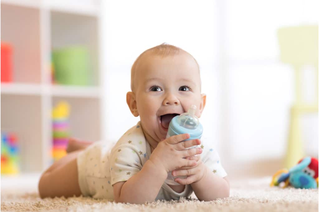 un bébé en body sur le ventre sur un tapis dans sa chambre, tient un biberon entre ses mains et le colle contre sa bouche grande ouverte.