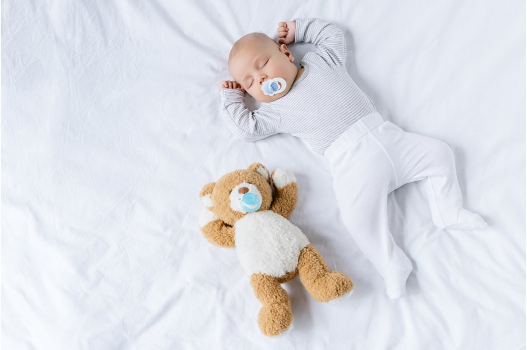 un bébé est endormi sur un lit tout blanc avec une tétine dans la bouche et un ourson en peluche à côté de lui avec une tétine dans sa bouche.