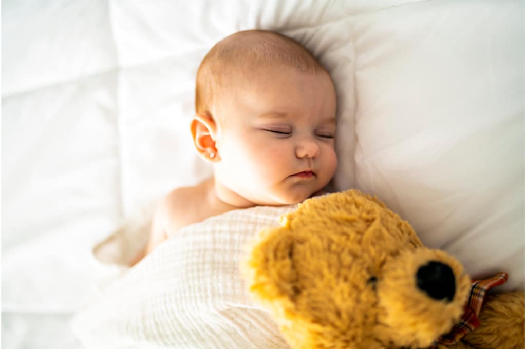 sur un lit, un bébé est endormi enveloppé d'un tissu blanc et aux côtés d'un ours en peluche.