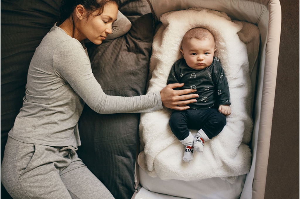 vue de dessus sur une maman en pyjama gris allongée sur le lit près de son bébé dans son berceau de cododo. le bébé ne dort pas, il est habillé, posé sur une couverture dans son berceau et regarde le photographe.