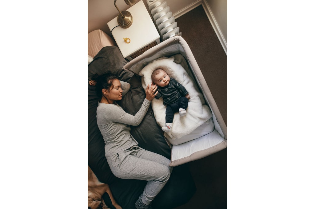 vue de dessus sur une maman en pyjama gris allongée sur le lit près de son bébé dans son berceau de cododo. le bébé ne dort pas, il est habillé, posé sur une couverture dans son berceau et regarde le photographe. une tétine est posé sur la table de chevet blanche.