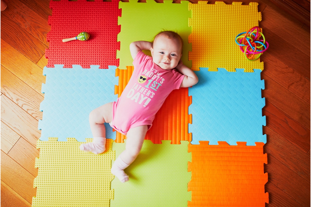 un bébé regarde avec le sourire le photographe. il est allongé sur un tapis de jeu posé au sol sur un parquet en bois. 2 jeux sont près du bébé.